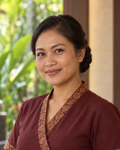 Ni Made Sari Dewi, Ploenjai therapist, smiling in the burgundy and gold uniform beside the garden fountain.